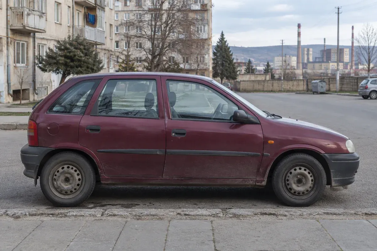 Бордовый старый хэтчбек на городской стоянке у жилых домов и промышленных труб